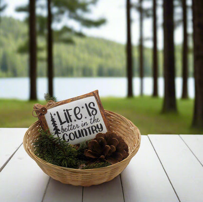 Decorative sign with 'Life is better in the country' text in a basket with greenery on a wooden table by a lake.