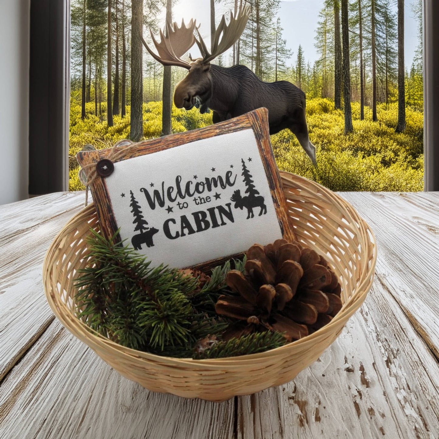 Decorative basket with 'Welcome to the Cabin' sign, pinecones, and evergreen branches in front of a forest background.