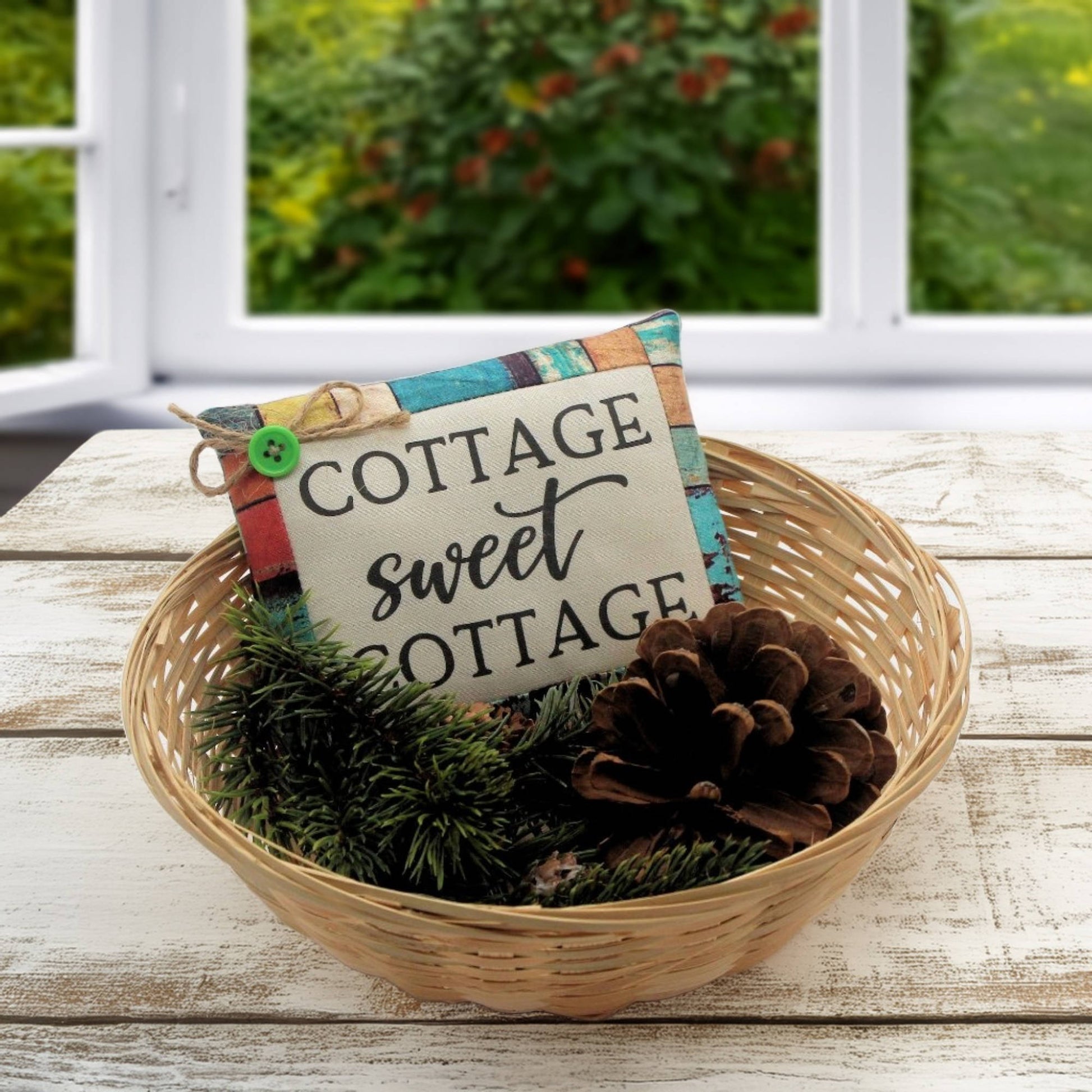 Decorative basket with 'Cottage sweet cottage' sign, pinecones, and greenery on a wooden surface.