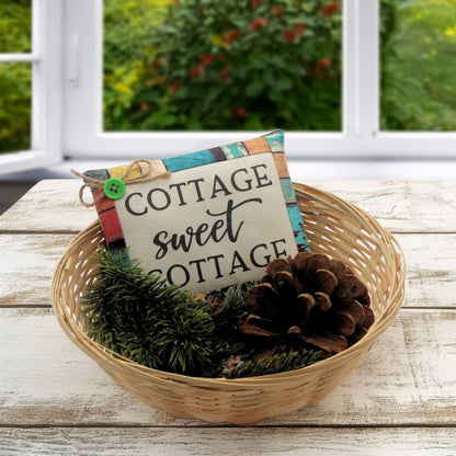 Decorative basket with 'Cottage sweet cottage' sign, pinecones, and greenery on a wooden surface.