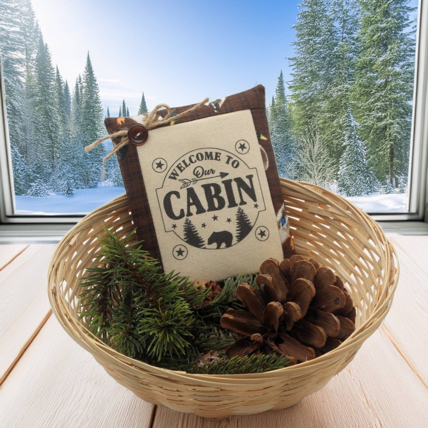 Decorative basket with 'Welcome to Our Cabin' sign and pinecones, set against a snowy cabin window backdrop.