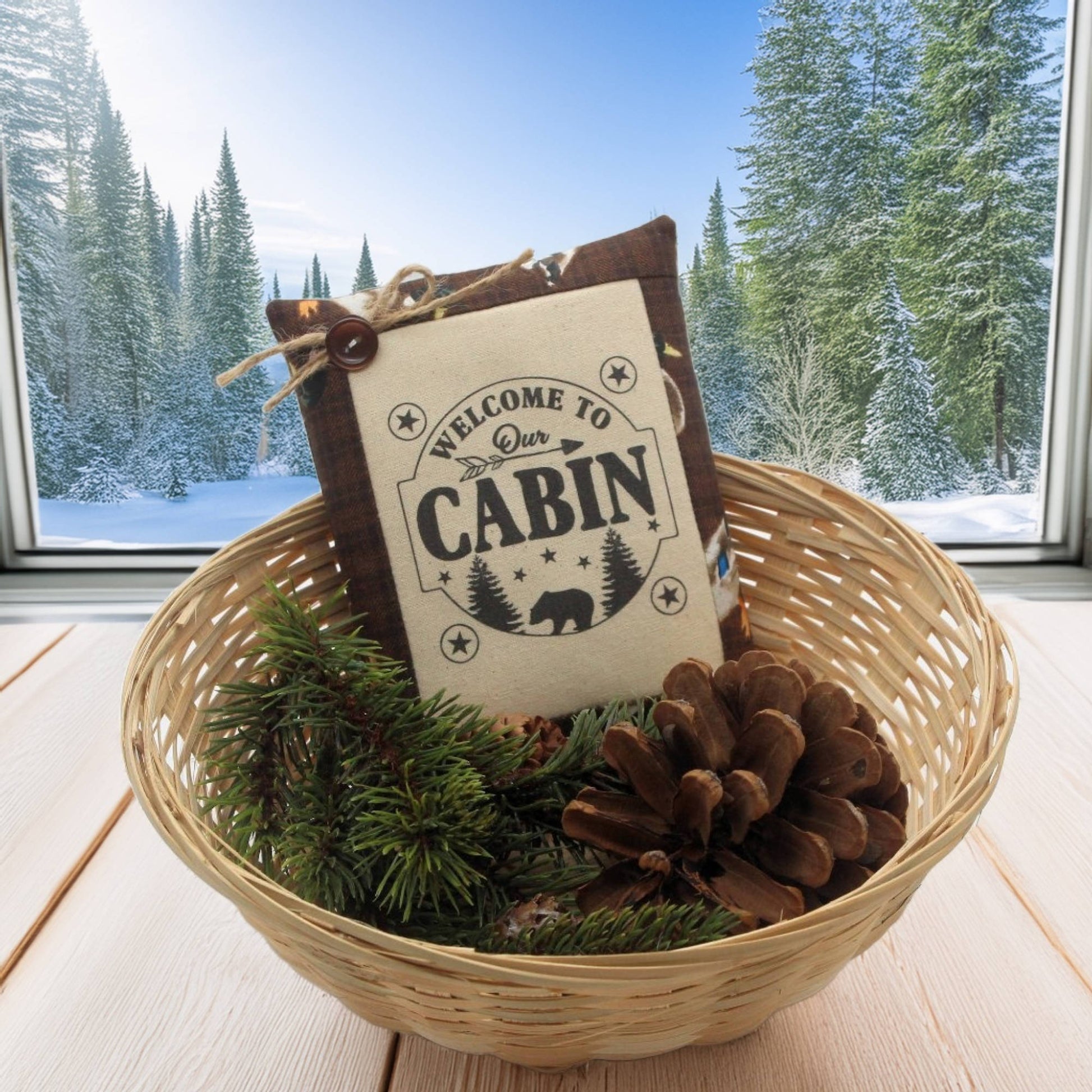 Decorative basket with 'Welcome to Our Cabin' sign and pinecones, set against a snowy cabin window backdrop.