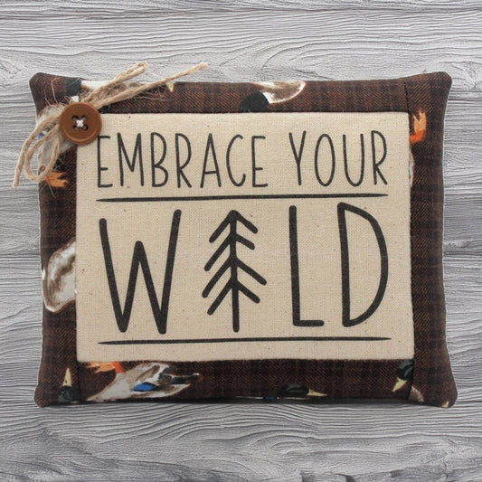 Decorative pillow tuck with 'Embrace Your Wild' text and tree symbol on a wooden surface