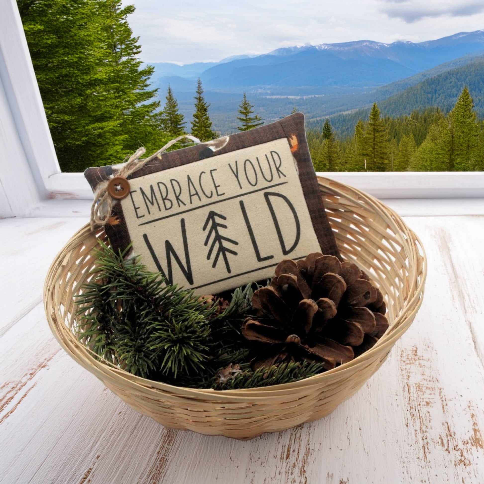 Decorative basket with pinecones, greenery, and a sign reading 'Embrace Your Wild' against a mountainous landscape.