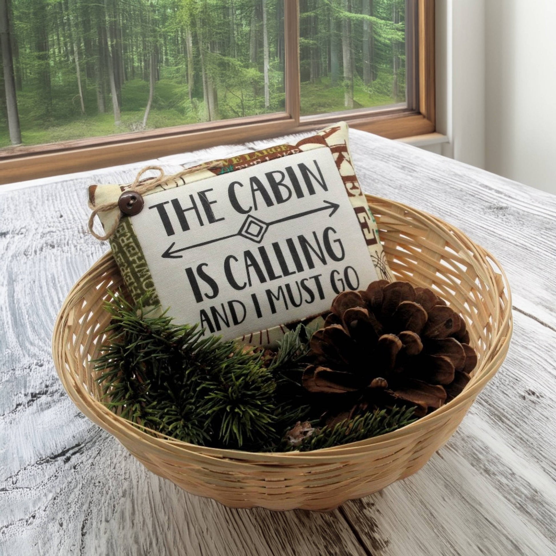 Decorative basket with a sign and pinecones on a wooden surface, with a forest view outside.