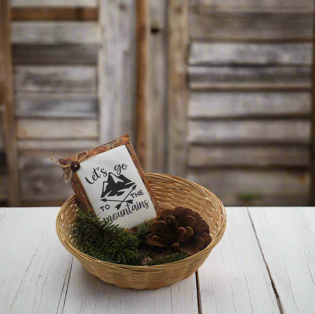 Decorative sign with 'Let's go to the mountains' text in a woven basket with pinecones on a wooden surface.