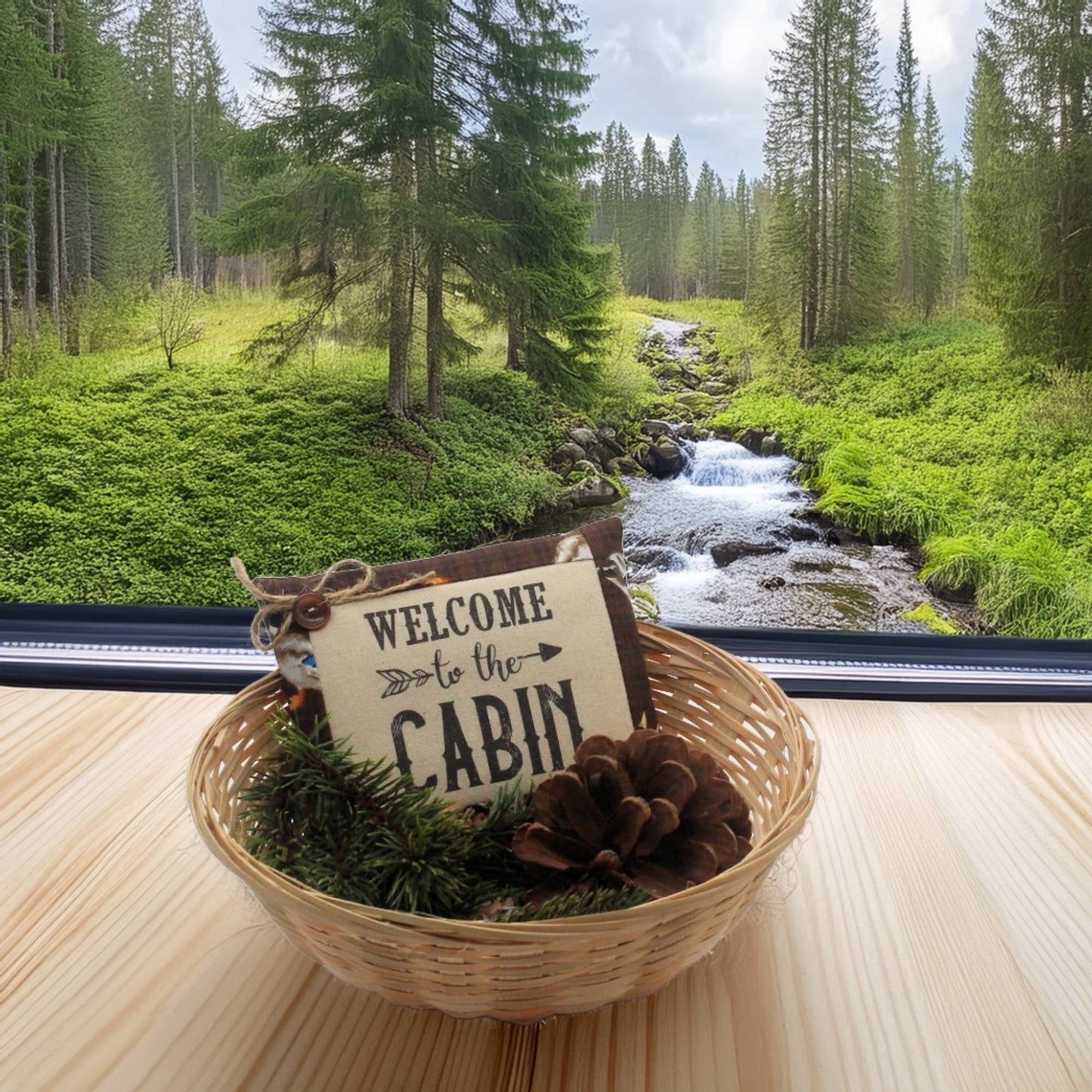 Decorative sign with 'Welcome to the Cabin' in a woven basket with pinecones and greenery, set against a forest backdrop.