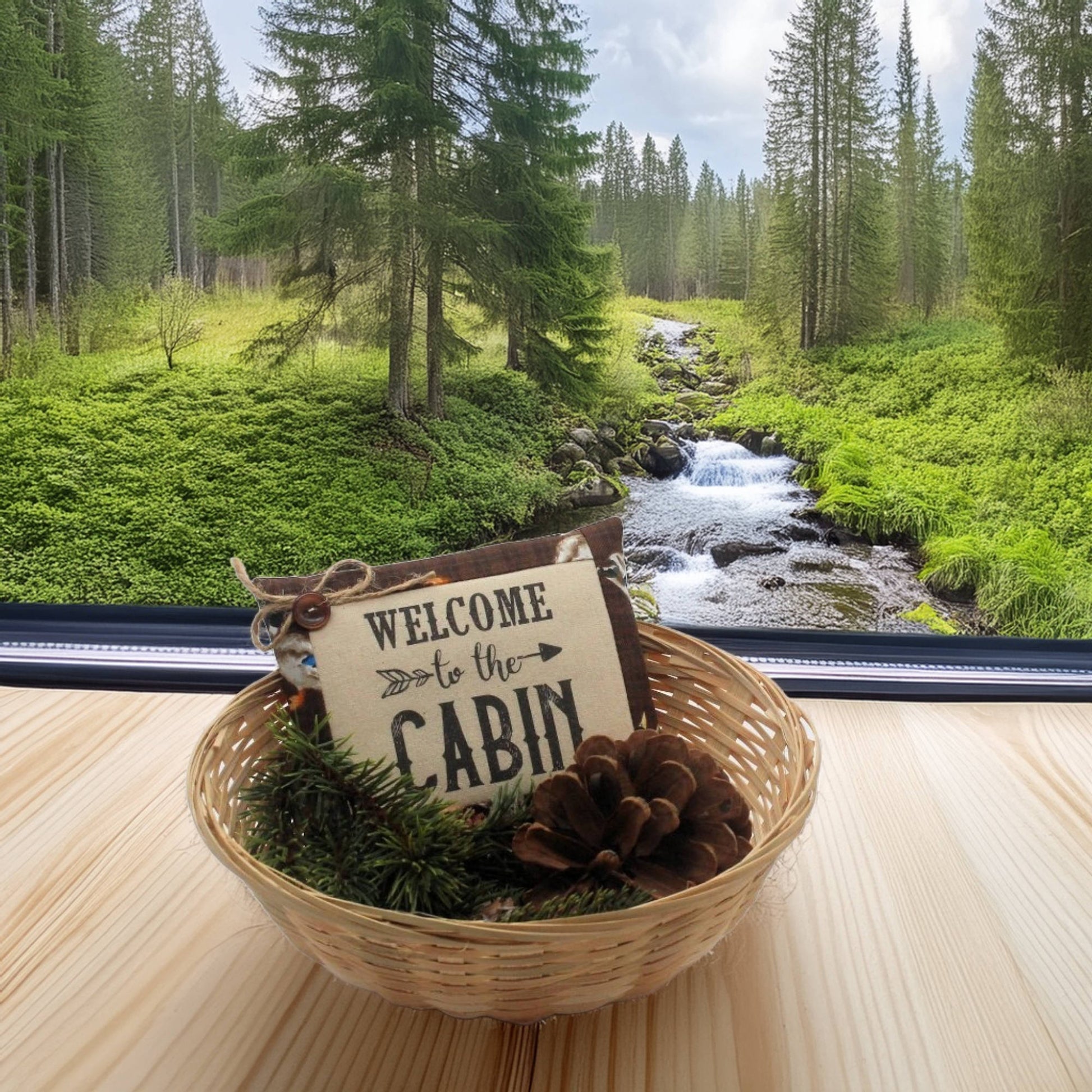 Decorative sign with 'Welcome to the Cabin' in a woven basket with pinecones and greenery, set against a forest backdrop.