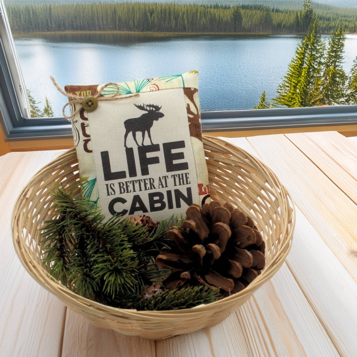 Decorative mini pillow with 'Life is Better at the Cabin' text in a basket with pine branches and pinecones, set against a scenic window view.