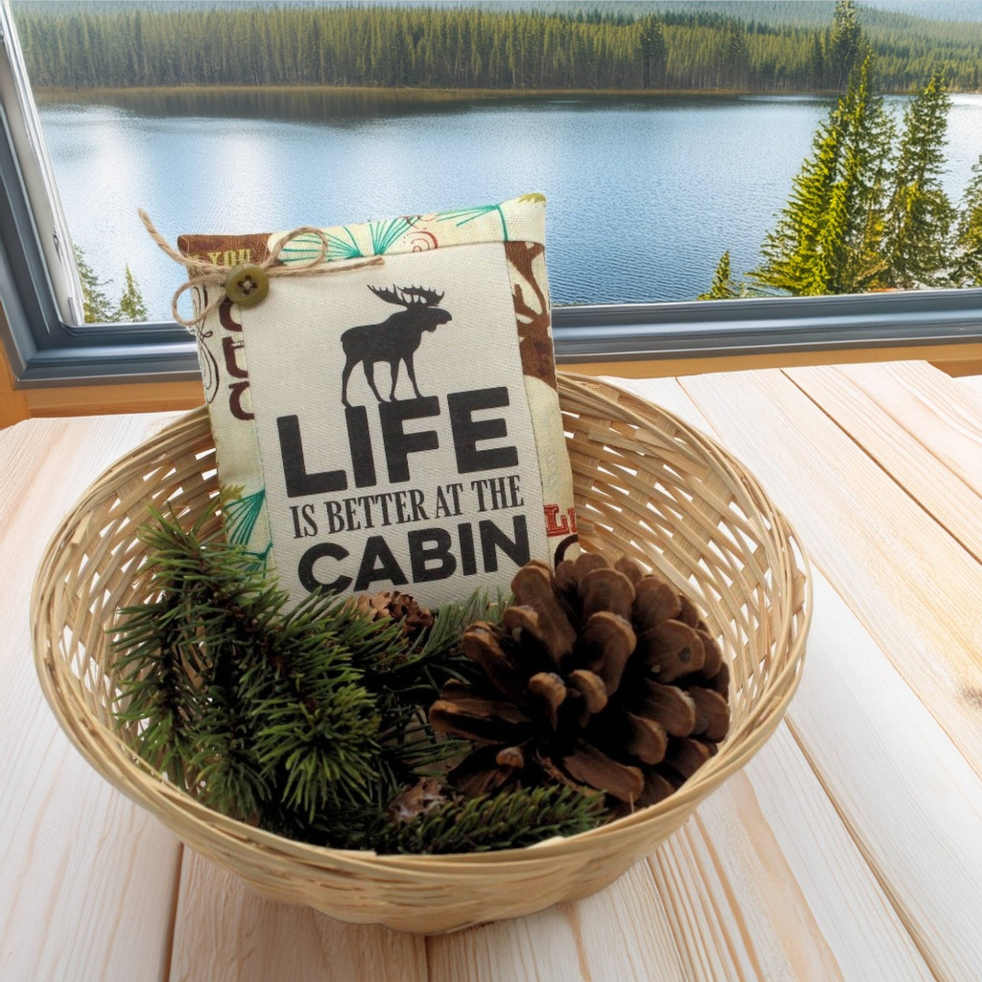 Decorative mini pillow with 'Life is Better at the Cabin' text in a basket with pine branches and pinecones, set against a scenic window view.