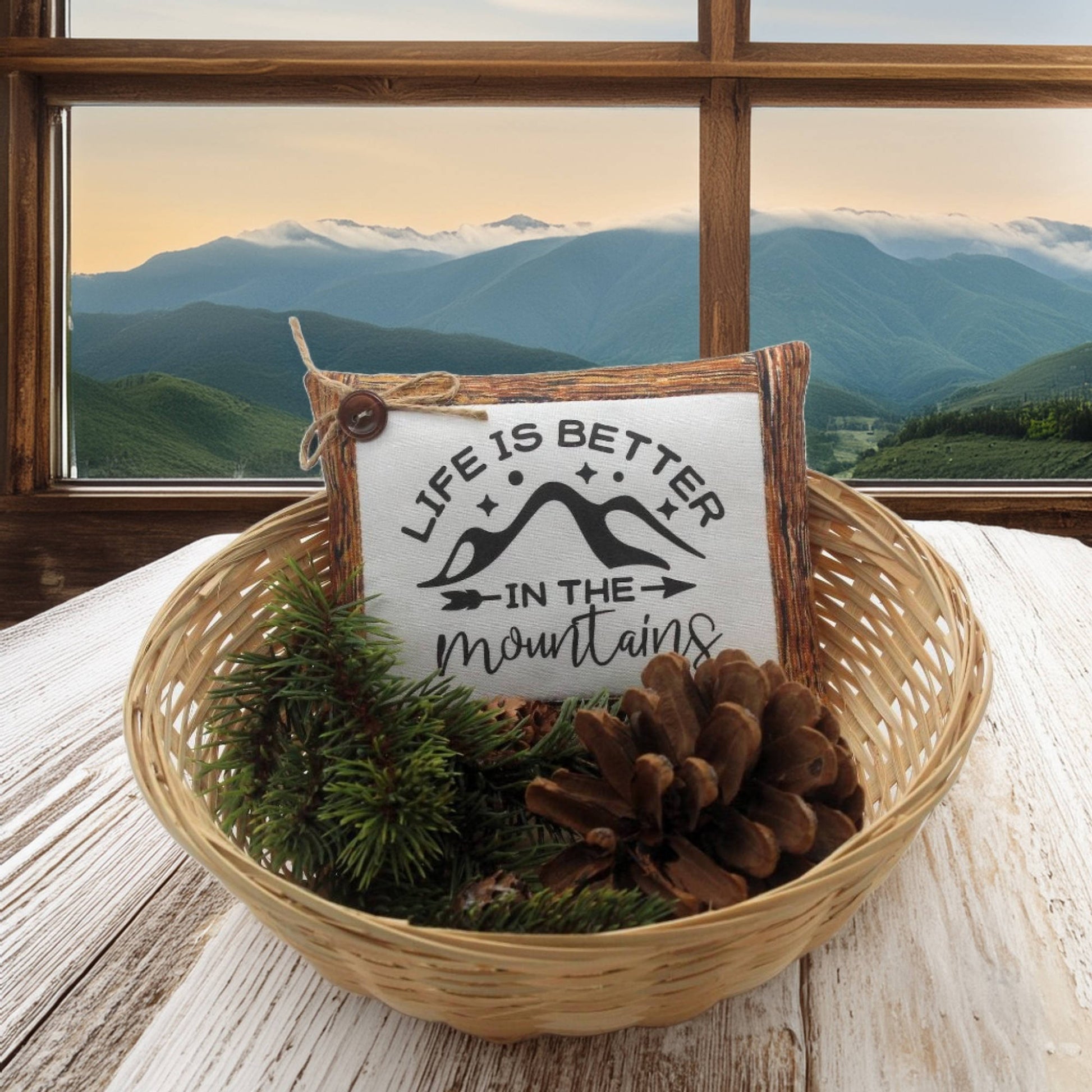 Decorative pillow with 'Life is better in the mountains' text in a woven basket with pinecones and greenery, set against a mountainous landscape.