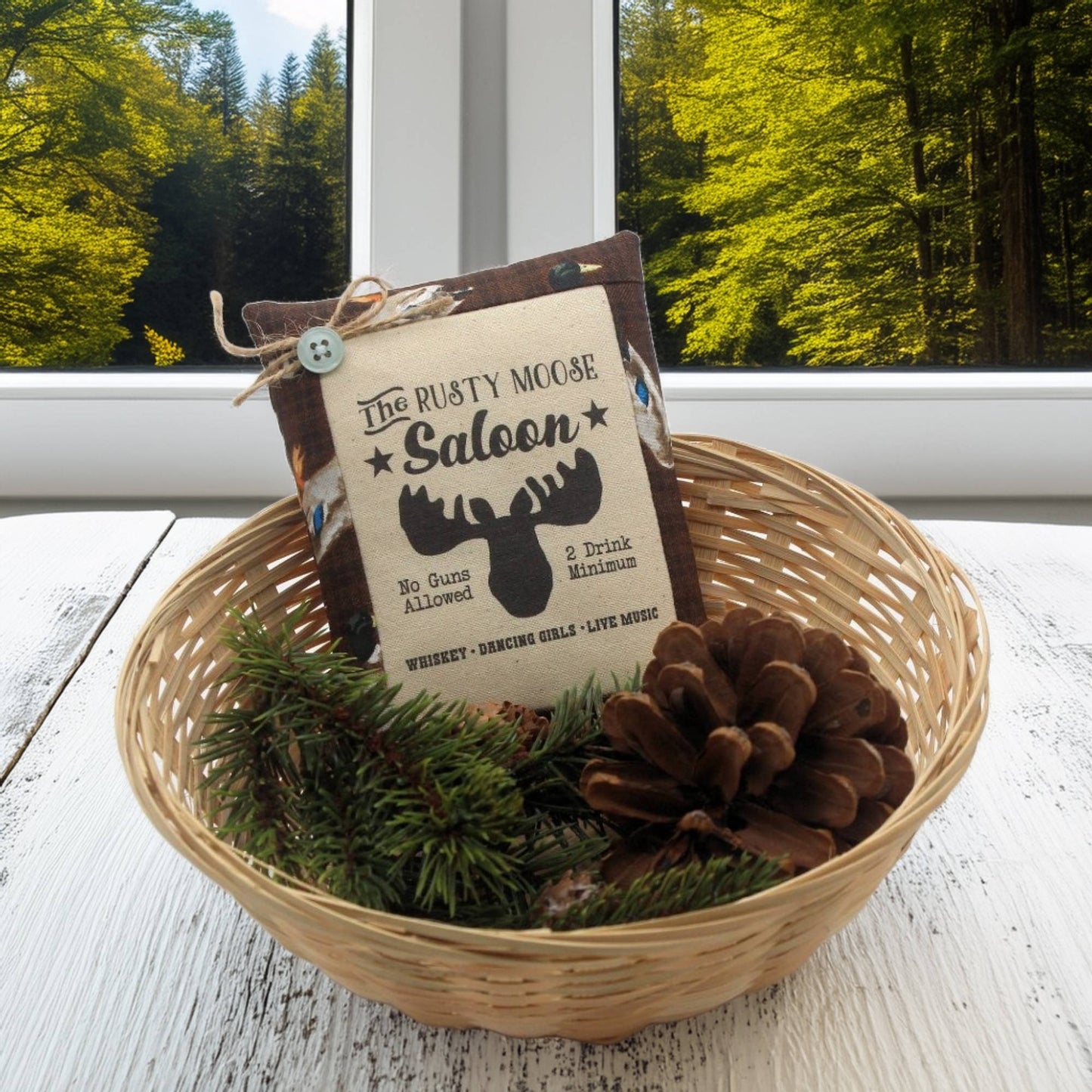 Decorative basket with a sign and pinecones on a wooden surface with a window view of trees.