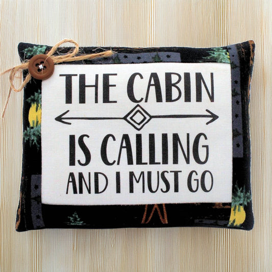 Decorative mini pillow with text 'The cabin is calling and I must go' on a wooden background