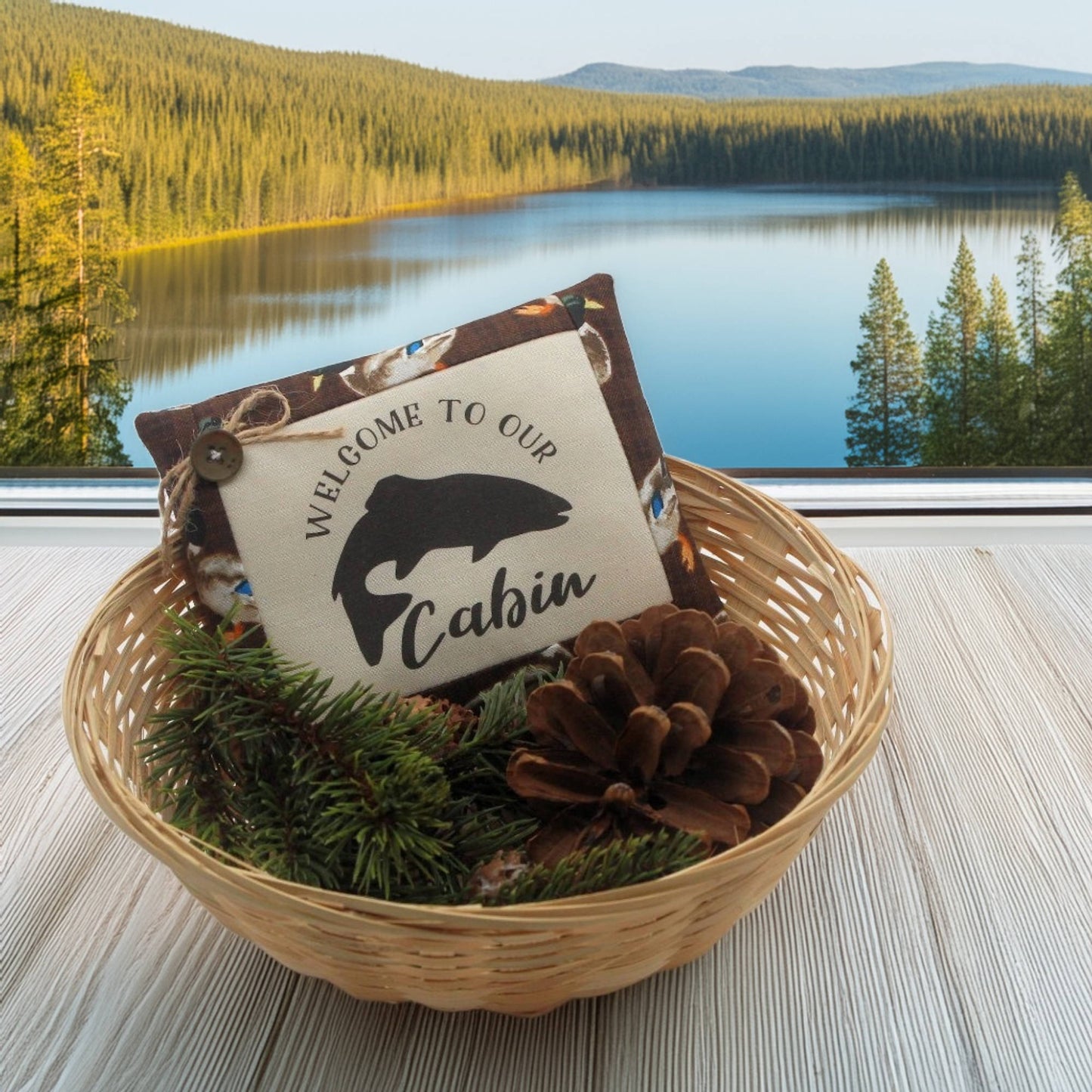 Decorative pillow with 'Welcome to our Cabin' sign in a woven basket with pinecones and evergreen branches, set against a scenic lake and forest background.