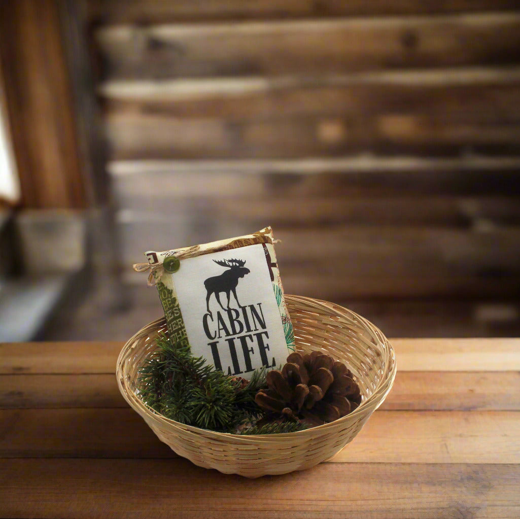 Decorative sign with 'Cabin Life' text and moose graphic in a woven basket on a wooden surface.