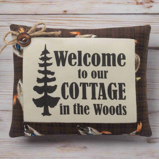 Decorative pillow tuck with 'Welcome to our Cottage in the Woods' text and tree design on a wooden background