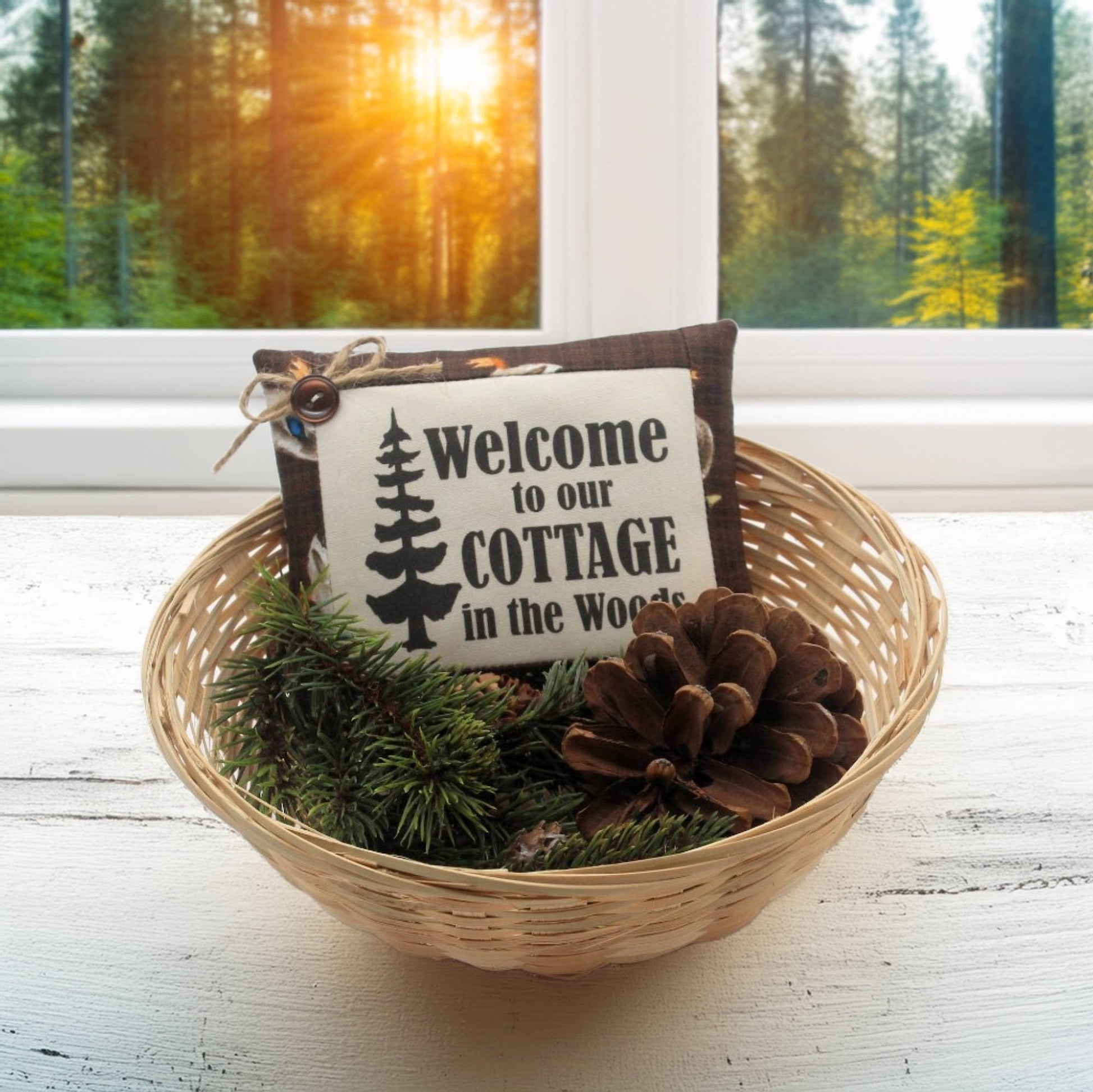 Decorative basket with pinecones and a 'Welcome to our Cottage in the Woods' sign on a windowsill with a forest view.