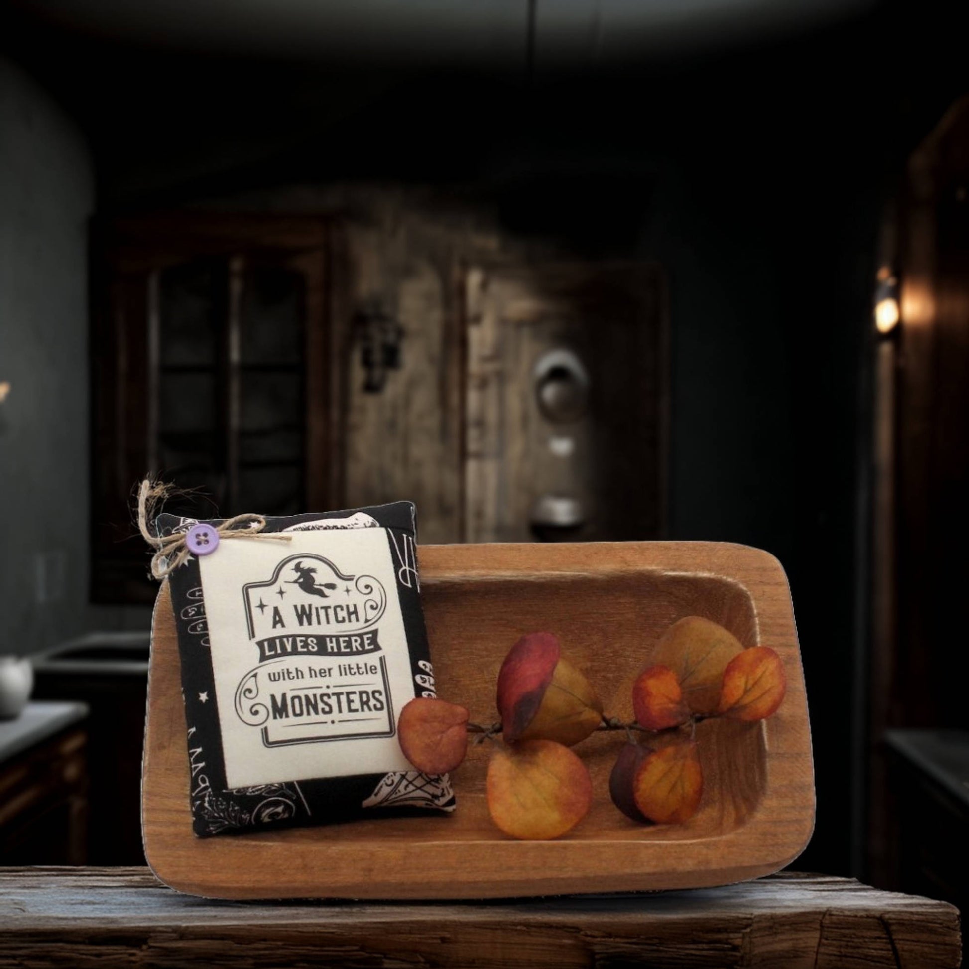 Wooden bowl with decorative foliage and a sign reading 'A Witch Lives Here with her Little Monsters' in a dark, rustic setting.