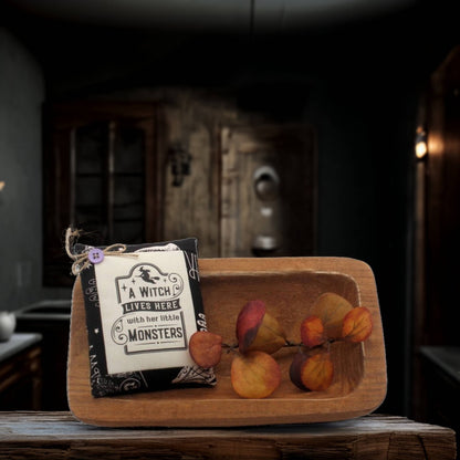 Wooden bowl with decorative foliage and a sign reading 'A Witch Lives Here with her Little Monsters' in a dark, rustic setting.