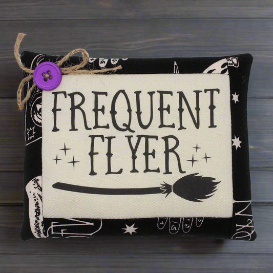 Decorative mini pillow with 'Frequent Flyer' text and broom graphic on a wooden background