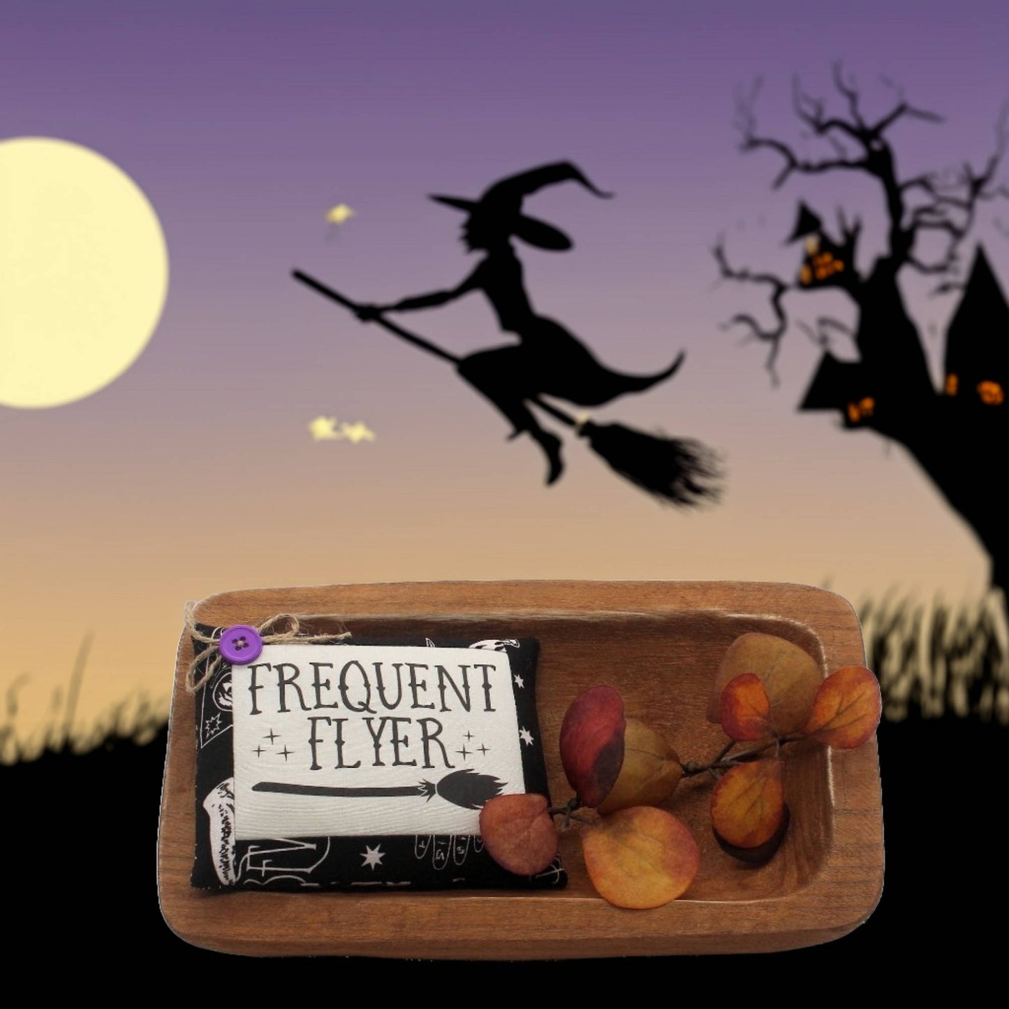 Wooden tray with 'Frequent Flyer' mini pillos against a Halloween-themed background with a witch flying.