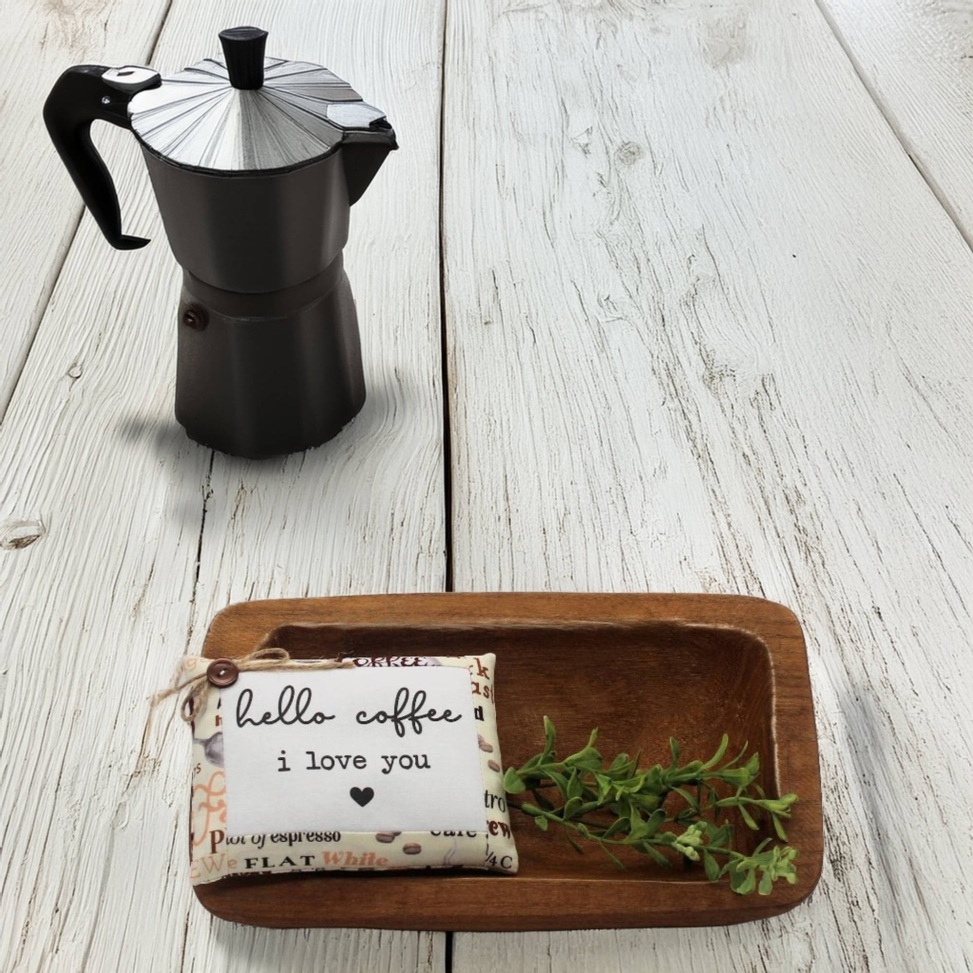 Black espresso maker on a wooden surface with a small wooden tray holding a pillow tuck and greenery.