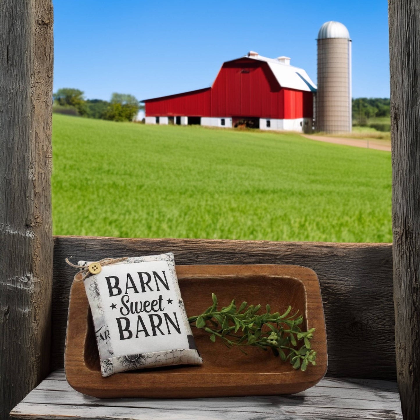 Decorative pillow with 'Barn Sweet Barn' text on a wooden surface with a red barn and silo in the background.