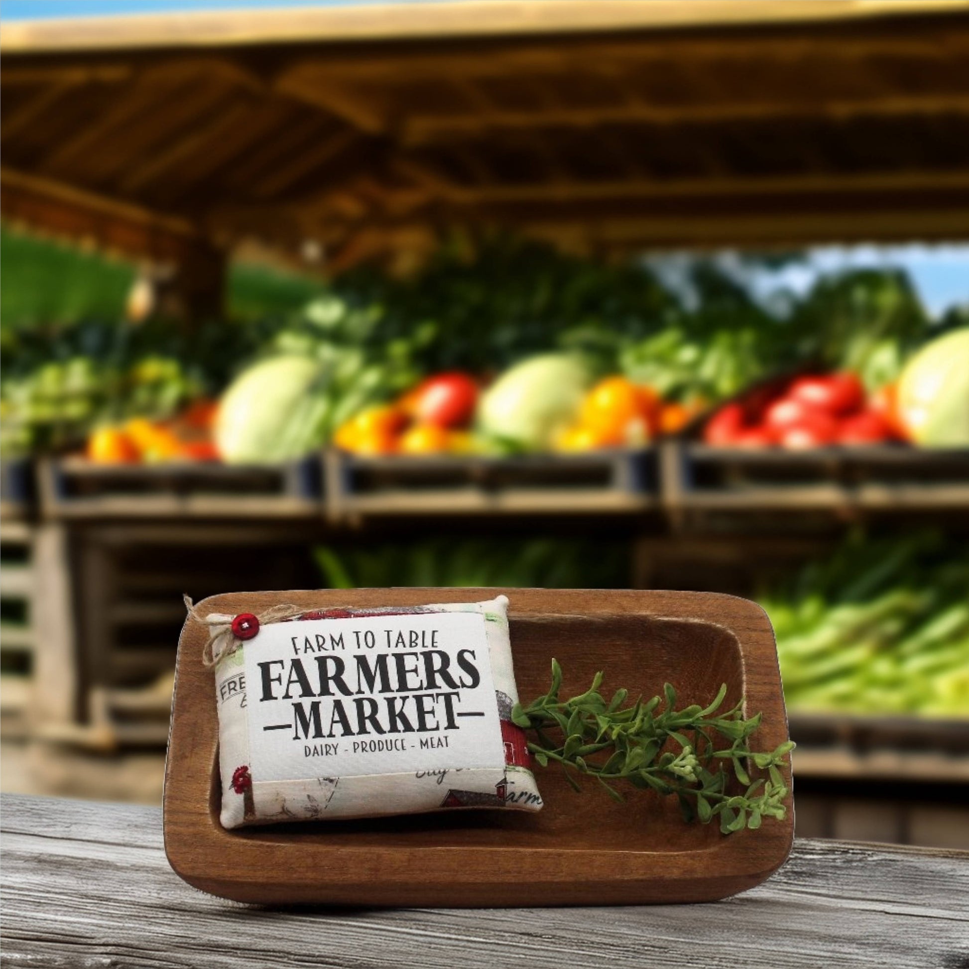 Wooden cutting board with a 'Farmers Market' product label and a sprig of greens on a wooden surface with a blurred background of vegetables.