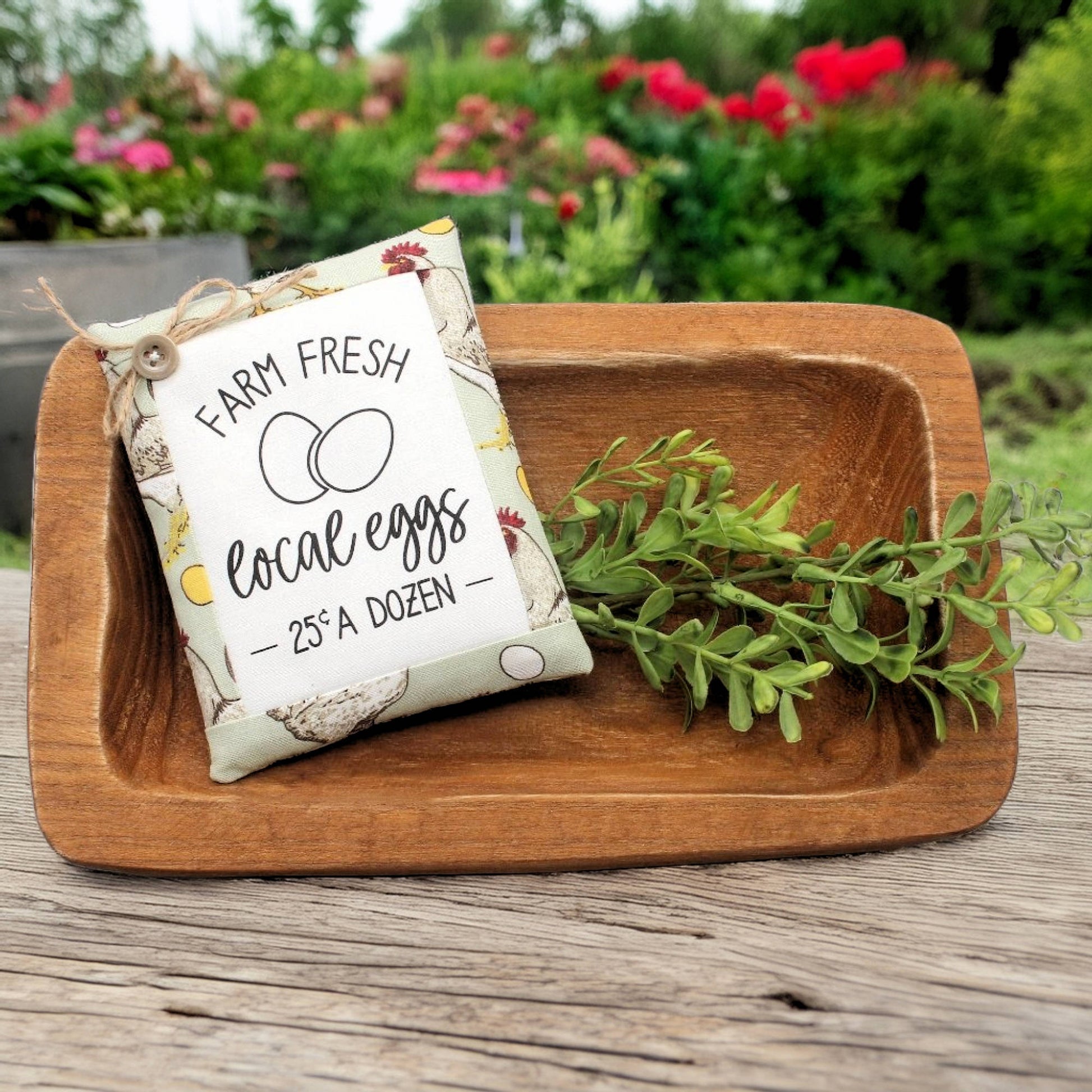 Wooden tray with a 'Farm Fresh Local Eggs' sign and greenery on a garden background
