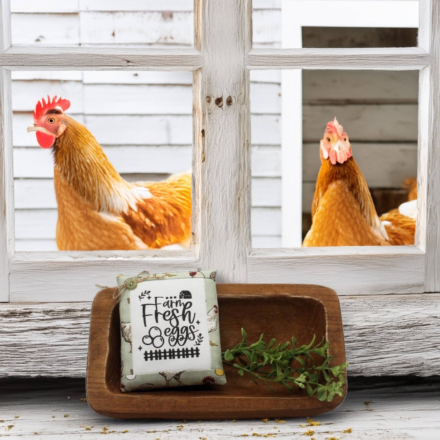 Two chickens peering through a window with a wooden tray holding a sign and greenery in front.