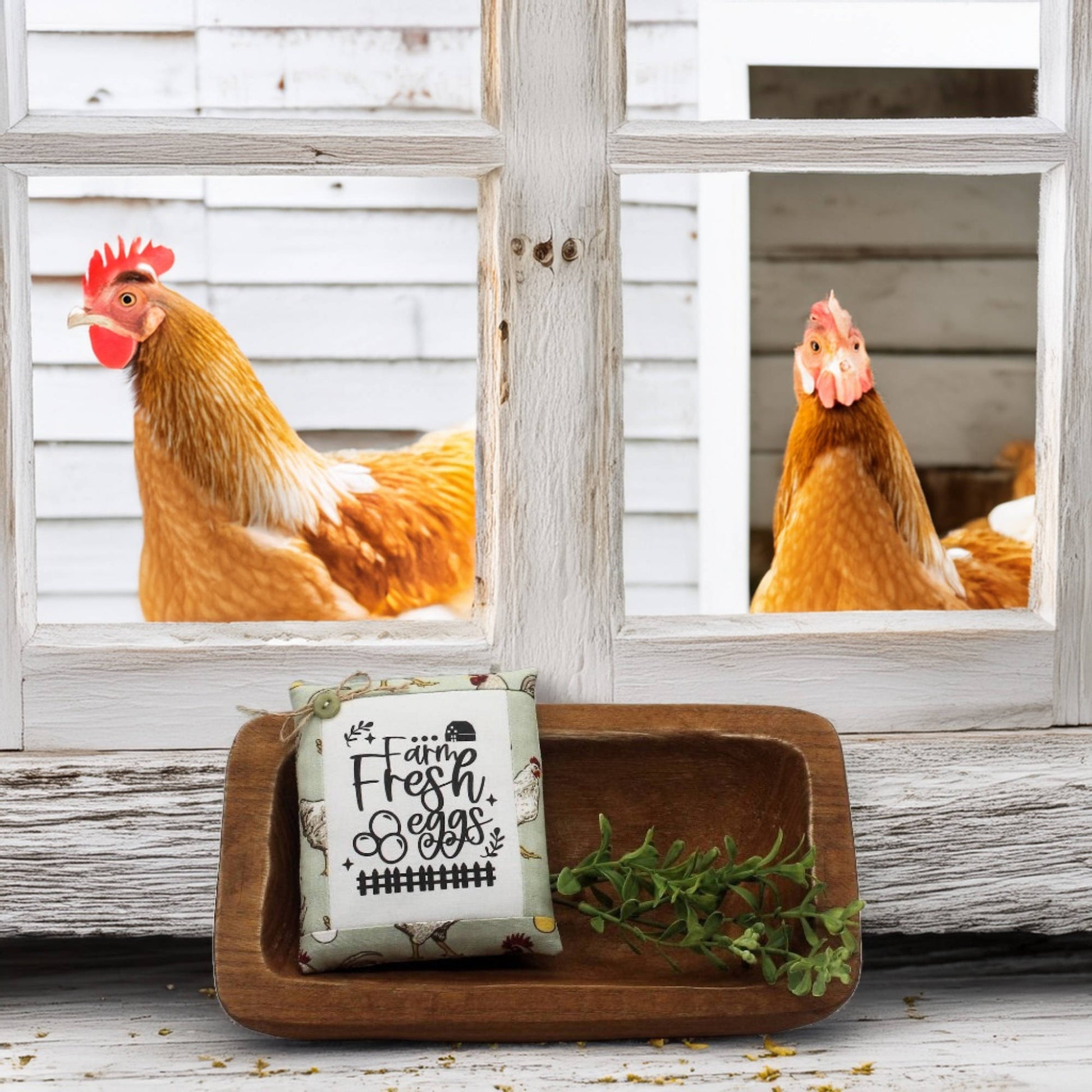 Two chickens peering through a window with a wooden tray holding a sign and greenery in front.