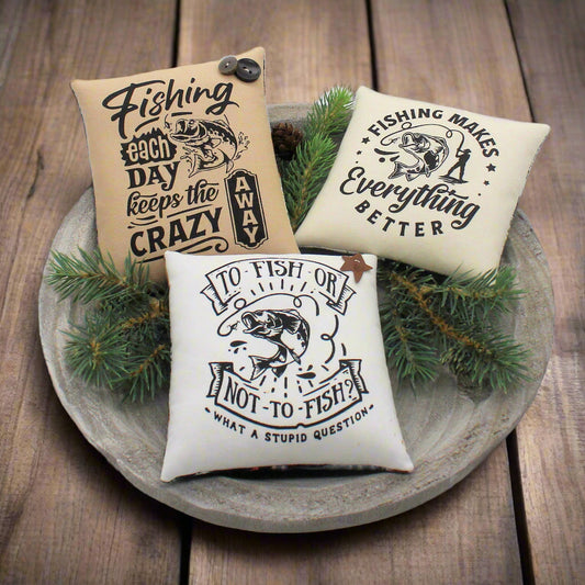 Three decorative pillows with fishing-themed sayings on a wooden tray with greenery.