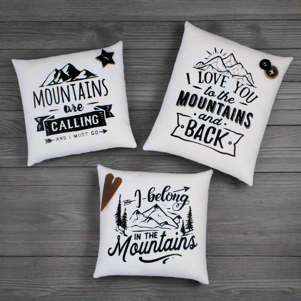 Three white pillows with mountain-themed text and graphics on a wooden surface