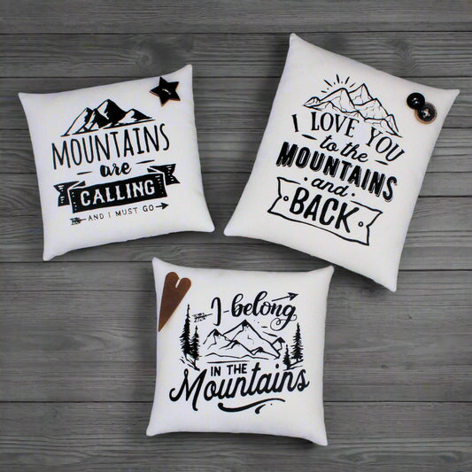 Three white pillows with mountain-themed text and graphics on a wooden surface