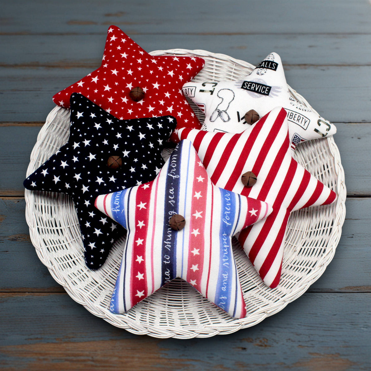 Five patriotic-themed star-shaped bowl fillers arranged on a round white wicker tray over a rustic blue-gray wooden surface. The pillows feature red, white, and blue patterns including stars and stripes, with rusty bells at the center of each star.