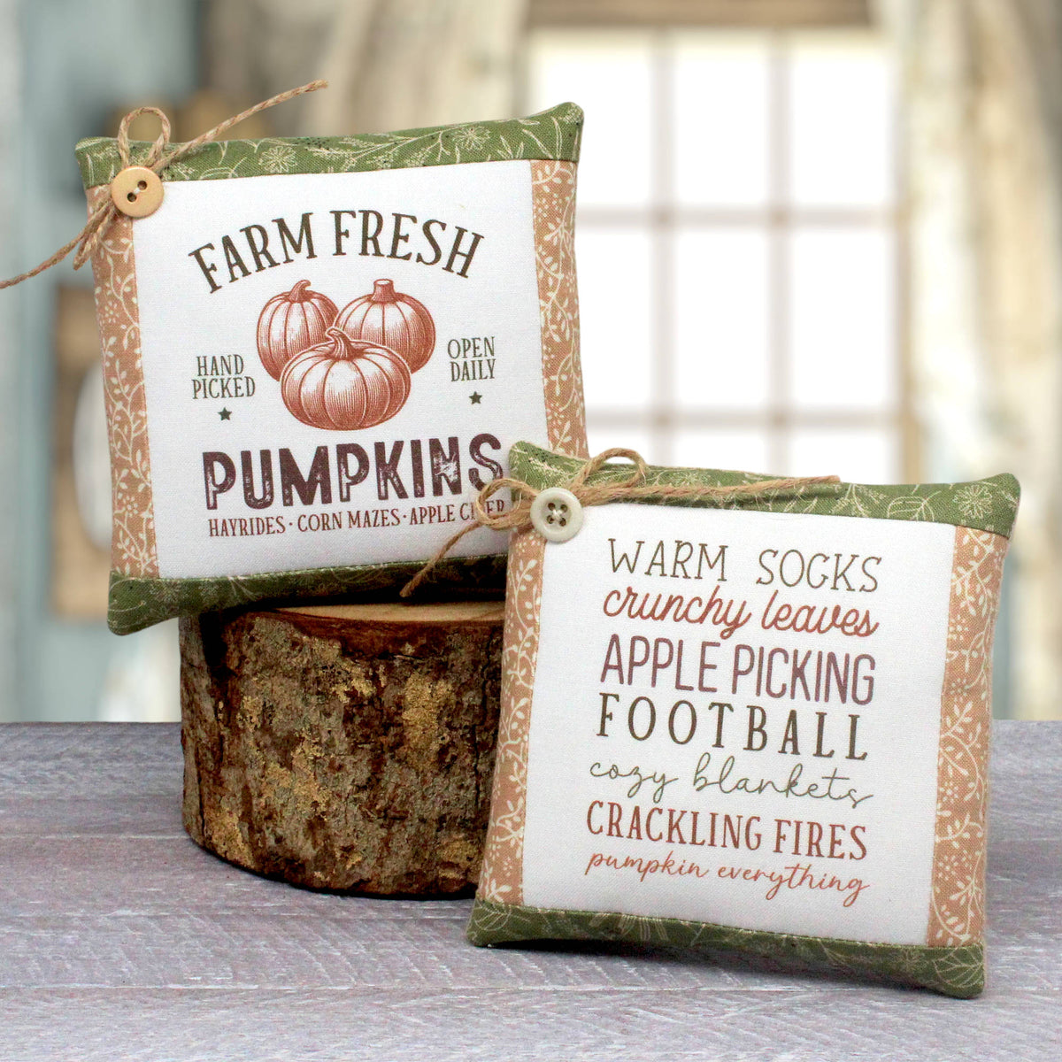 Two decorative fabric pillows with autumn-themed printed text and pumpkin graphics, tied with twine and buttons, displayed on a rustic wood slice with a softly blurred indoor background.