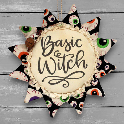 A sunflower-shaped fabric hanging ornament with the words “Basic Witch” in black script on a cream center, surrounded by pointed petals printed with multicolored eyeballs on a black background. The ornament is photographed against a gray wood plank surface.