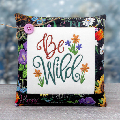 Small pillow tuck or bowl filler embroidered with 'be wild' and wildflowers, trimmed with a fabric that has a floral design on a black background.