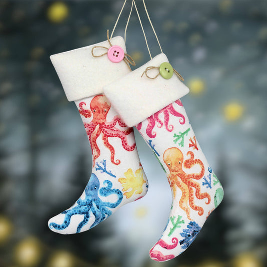 Two Christmas stockings hanging by twine, decorated with colorful octopus and sea plant illustrations, set against a dark blurred holiday background.