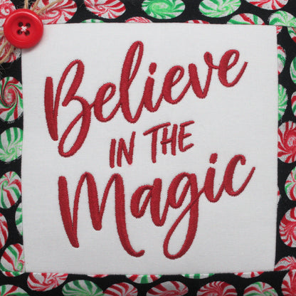 A square white fabric bowl filler pillow with red embroidered text reading 'Believe in the Magic,' shown on a black background with a red button and a candy cane pattern.
