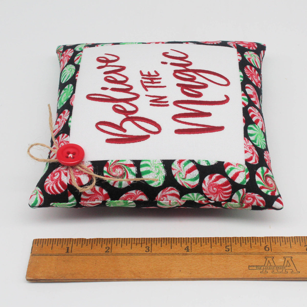 A small square fabric pillow with a white center panel reading 'Believe in the Magic' in red script, surrounded by a black border printed with red and green peppermint candies. A red button and twine are attached on the left side, and a wooden ruler is placed below the pillow for scale on a white background.