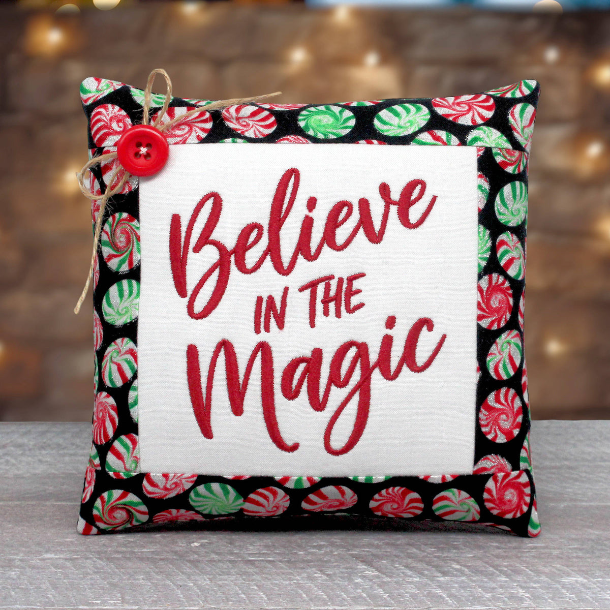 A square decorative throw pillow with a festive holiday design. The center is a white fabric panel with red embroidered text reading 'Believe IN THE Magic'.