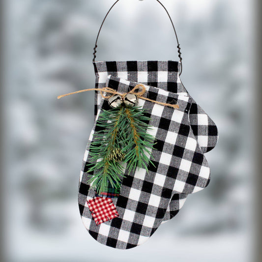 A Christmas mittens ornament made from black and white buffalo plaid fabric, decorated with a green pine branch, small silver bells, a twine bow, and a red and white gingham accent patch, hanging against a blurred snowy background.