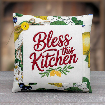 A square decorative throw pillow with a white center panel featuring the embroidered phrase 'Bless this Kitchen' in red script, surrounded by a printed border with green leaves, yellow lemons, and blue berries. A small yellow button and twine are attached near the top left.