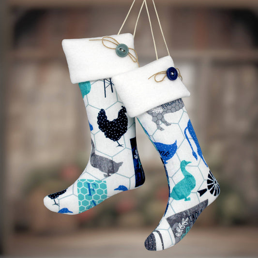 Two white fabric Christmas stockings with a blue, teal, and gray farm animal and chicken wire pattern, hanging against a blurred rustic background.