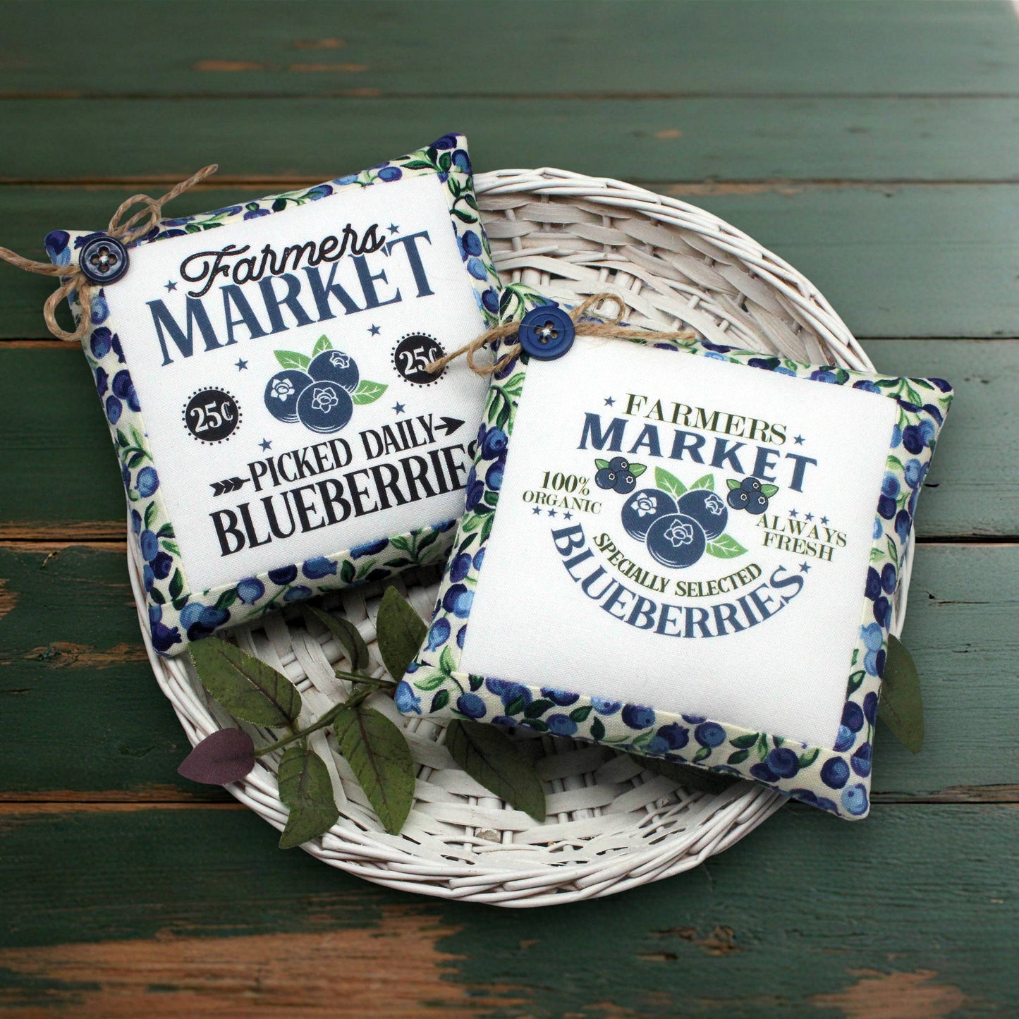 Two decorative square fabric mini pillows with a blueberry-themed 'Farmers Market' print, arranged on a white wicker tray over a distressed green wooden surface.