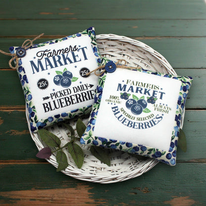 Two decorative square fabric mini pillows with a blueberry-themed 'Farmers Market' print, arranged on a white wicker tray over a distressed green wooden surface.
