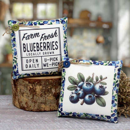 Two decorative square fabric pillows with blueberry-themed designs, displayed on a rustic wood slice and tabletop in a softly blurred indoor setting. One pillow has a white front with black text reading 'Farm Fresh Blueberries Locally Grown' and a blueberry-patterned border.