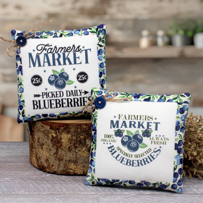 Two decorative fabric mini pillow-like signs with a blueberry-themed 'Farmers Market' design, displayed on a rustic wood stump and tabletop in a styled indoor setting.