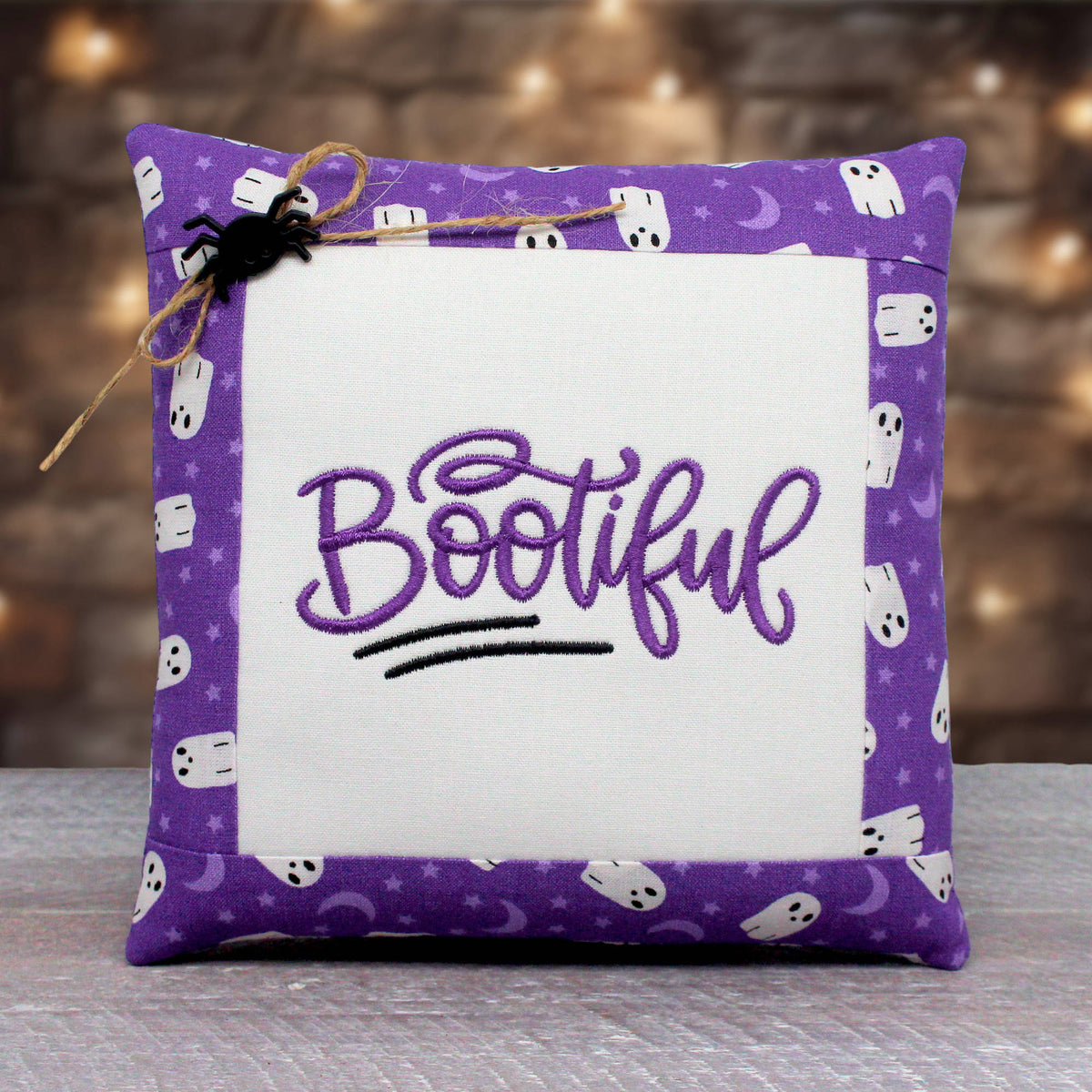 A square decorative throw pillow with a purple border patterned with white ghosts, moons, and stars, featuring a white center panel with the word 'Bootiful' embroidered in purple script. A small black spider decoration and twine are attached to the top left corner.